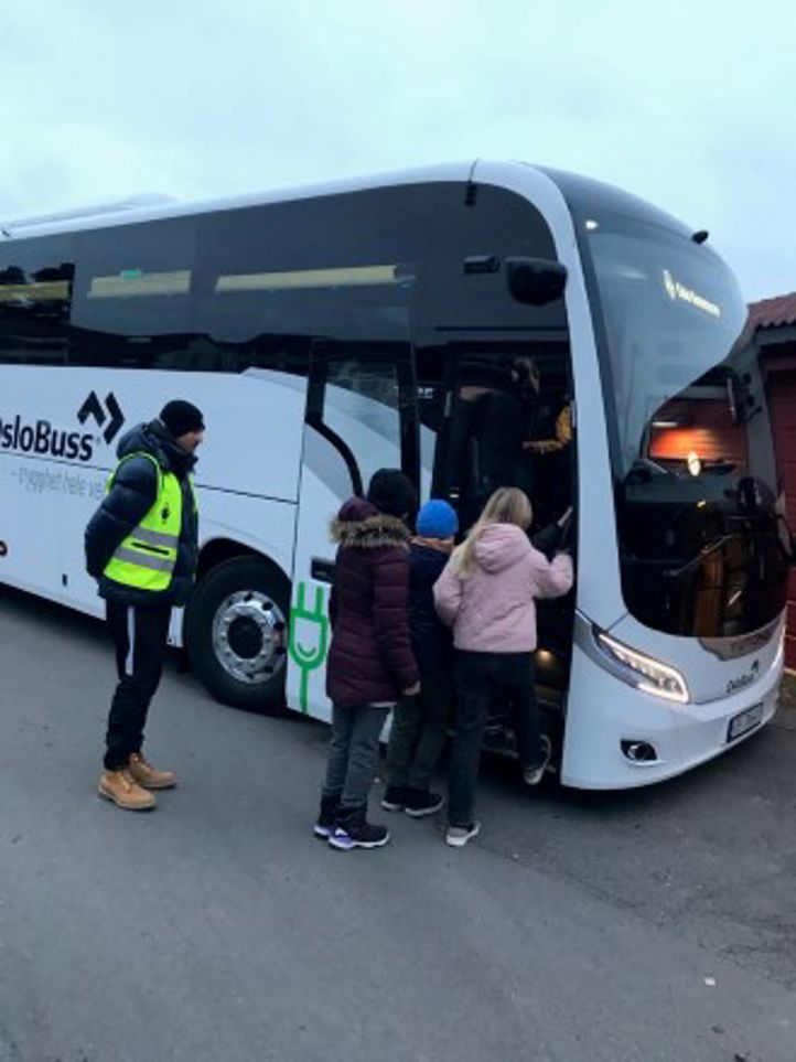 Foto: Indra Dart En hvit buss står ved skolen og elever er på vei inn i bussen.