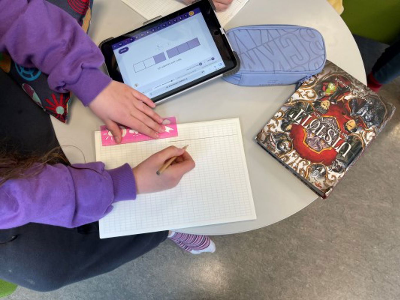 På et lite rundt bord ligger det en Ipad, et pennal og en skrivebok som eleven gjør matteleksen i.